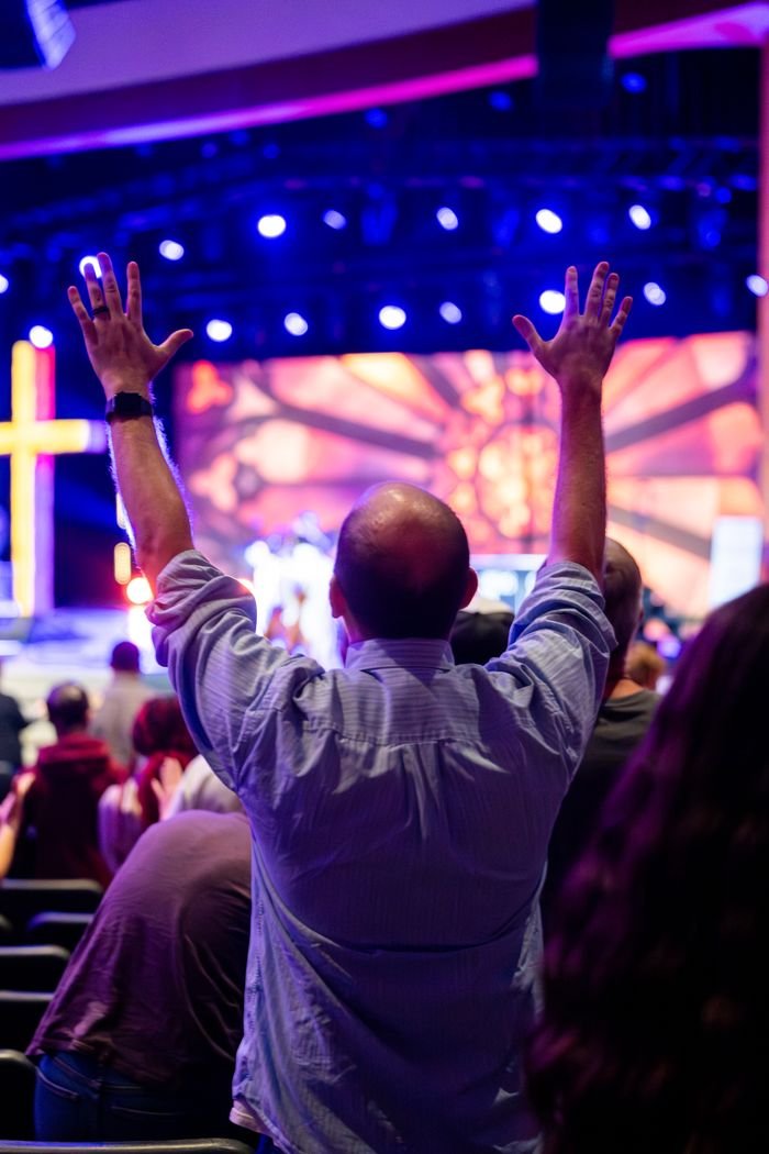 Congregation member raising hands in worship during a lively church service with colorful lights and cross.